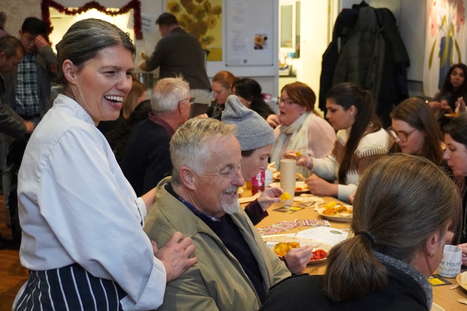 A smiling woman standing with her hand on a seated man's shoulder, surrounded by others eating at a long table.