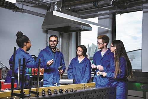 Five engineering researchers in an industrial laboratory wearing blue boiler suits