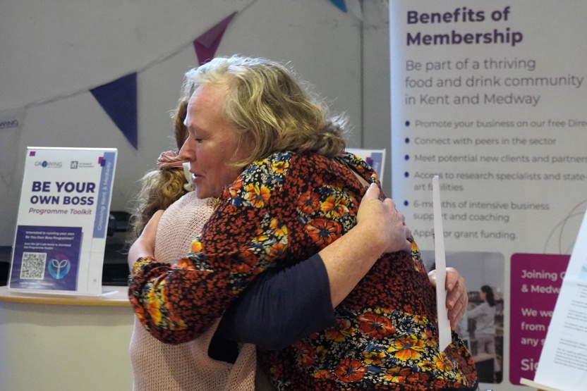 Two women hugging in front of Growing Kent & Medway's display at Sunflower House.